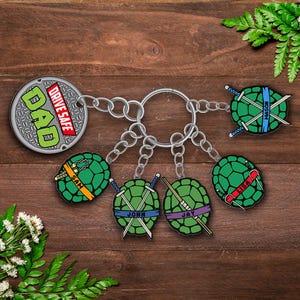 May include: A set of keyrings featuring a manhole cover with "DAD DRIVE SAFE" text and turtle shell designs with names. The keyrings are attached to a large metal ring. The background is a wooden surface.