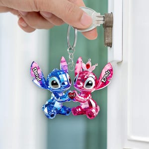 May include: A keychain with two crystal-effect figures, one blue and one pink, holding a heart with "Love." The blue figure has "David" on its ear, and the pink figure has "Emily." The figures are attached to a silver keyring.