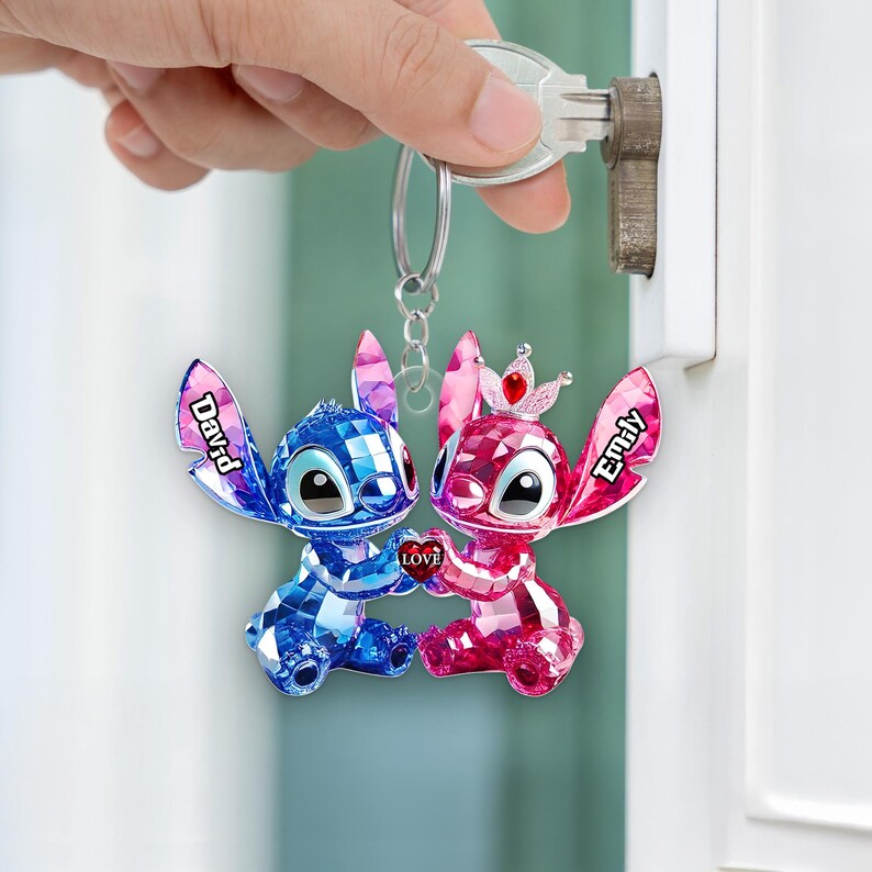 May include: A keychain with two crystal-effect figures, one blue and one pink, holding a heart with "Love." The blue figure has "David" on its ear, and the pink figure has "Emily." The figures are attached to a silver keyring.