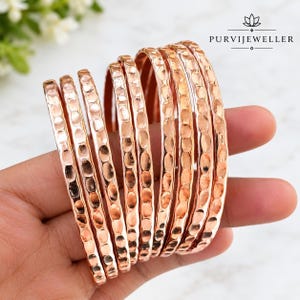 Può includere: Un set di otto bracciali rigidi color oro rosa con una superficie martellata e testurizzata. Ogni bracciale ha un profilo arrotondato e una finitura metallica riflettente. Il marchio PURVIJEWELLER è stampato in nero nell'angolo in alto a destra.