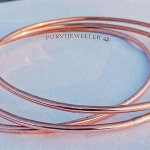 Può includere: Tre bracciali rigidi circolari sottili color rame con una finitura liscia e lucida. I bracciali sono impilati sovrapponendosi leggermente. Il testo PURVIJEWELLER è stampato in piccole lettere scure vicino al centro superiore dei bracciali.