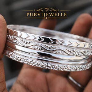 Pode incluir: Um conjunto de pulseiras de prata com designs intrincados, incluindo padrões de chevron, florais e ondulados. As joias são seguradas em uma mão, mostrando o metal polido e o artesanato detalhado. O nome da marca "PURVIJEWELLE" é exibido.