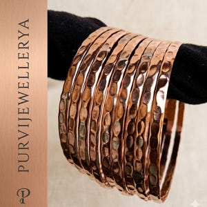 Peut inclure: Un ensemble de sept bracelets joncs de couleur cuivre présentant une surface martelée et texturée. Les bracelets en métal sont disposés en pile. Le texte PURVIJEWELLERYA est visible sur le côté gauche, avec un logo en forme de lettre P.