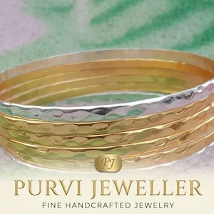 Peut inclure: Un empilement de cinq joncs fins avec une texture martelée. L'ensemble comprend des anneaux en métal alternant tons argentés et dorés. Une petite étiquette ovale avec les lettres PJ est fixée à l'avant. Le texte PURVI JEWELLER FINE HANDCRAFTED JEWELRY est imprimé sous les joncs.