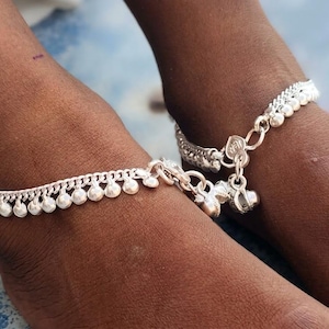 May include: A pair of silver-colored anklets featuring a delicate chain design adorned with small, round, dangling charms along the length. The anklets are secured with a metal clasp and include additional decorative bell-shaped charms at the closure.