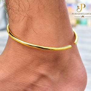 May include: A polished gold-tone anklet featuring a smooth, open-ended circular band design. The jewelry piece has a simple, continuous metallic finish that wraps around the ankle, showcasing a clean and reflective surface with rounded ends.