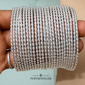 Può includere: Un set di ventiquattro braccialetti rigidi color argento, ognuno caratterizzato da una texture intrecciata simile a una corda. I braccialetti sono disposti in una pila, mostrando una finitura metallica uniforme e una forma circolare costante.