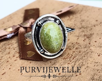 Könnte beinhalten: Ein Silberring mit einem ovalen, gesprenkelten hellgrünen Stein, der in einen polierten Metallrahmen mit kleinen dekorativen Kugeln gefasst ist. Der Text PURVIJEWELLE steht unter dem Ring, der auf einer strukturierten beigen Fläche liegt.