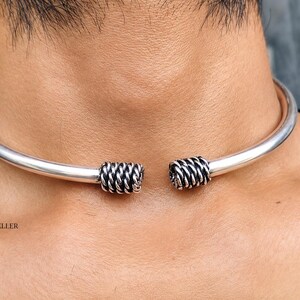Op de afbeelding: Een zilverkleurige open choker-halsketting met een gladde, gepolijste band die aan beide uiteinden eindigt in een getextureerd, touwachtig ontwerp. Het metalen oppervlak reflecteert het licht en de tekst PURVIJEWELLER is zichtbaar.
