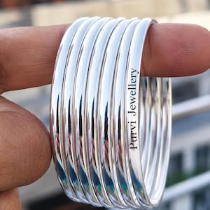 May include: A stack of shiny silver bangles. The bangles are smooth and reflective, with the text "Purvi Jewellery" engraved on one of the bangles. The bangles are held by a finger.