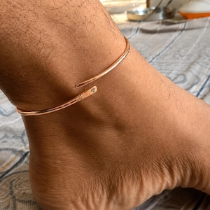 May include: A rose gold-toned metal anklet featuring a smooth, open-ended cuff design. The anklet has a polished finish and includes a small, engraved diamond-shaped symbol near one of the tapered ends, which rests against the skin.