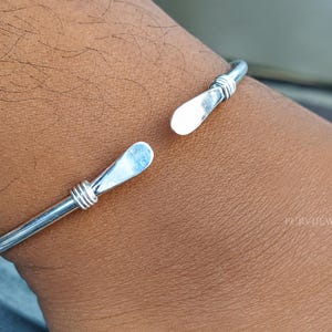 May include: A silver-toned open cuff bracelet featuring a smooth, polished finish. The bracelet has two flat, paddle-shaped ends, each accented with three thin, wrapped wire details just below the flattened terminals, creating a clean, metallic look.