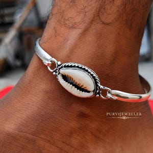May include: A silver-toned metal anklet featuring a central oval cowrie shell. The shell is framed by a twisted rope-style silver border and connected to a smooth, polished silver band. The text PURVIJEWELLER is visible near the bottom right.