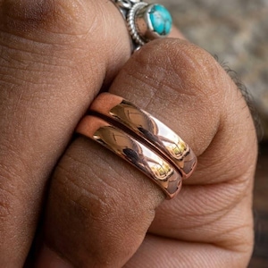 May include: Two polished copper rings with a smooth, rounded band design. The rings feature a warm, metallic reddish-brown finish that reflects light. A separate ring with a small, round, blue-green turquoise stone is visible on the adjacent finger.