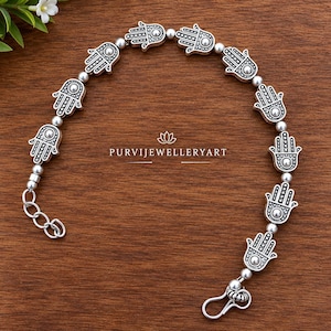 May include: A silver-toned bracelet featuring a series of Hamsa hand charms alternating with small round beads. The metal charms display intricate engraved details. The text PURVIJEWELLERYART is centered within the arc of the bracelet.