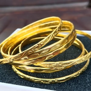 Könnte beinhalten: Ein Set aus vier goldfarbenen Armreifen mit polierter Oberfläche. Jeder Armreif weist ein sich wiederholendes geprägtes Blumenmuster und eine Reihe glatter, abgerundeter Vertiefungen auf der Außenfläche auf, was für eine Textur sorgt.