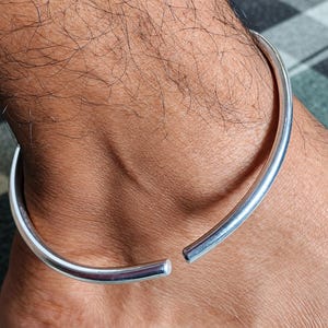May include: A polished silver-tone metal anklet featuring a simple, open-ended circular design with rounded tips. The band has a smooth, reflective surface and is worn around the ankle, showcasing a minimalist and continuous metallic construction.
