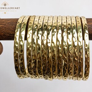 Op de afbeelding: Een set van twaalf goudkleurige metalen armbanden met een gehamerd, getextureerd oppervlak. Elke armband heeft een afgerond profiel en een reflecterende afwerking. De armbanden zijn op een rij gerangschikt en tonen het consistente putjespatroon.