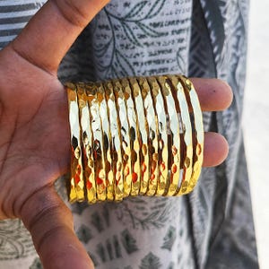 Op de afbeelding: Een verzameling goudkleurige armbanden met een gehamerde textuur. De armbanden zijn op elkaar gestapeld, reflecteren licht en creëren een glinsterend effect. De sieraden worden in een hand gehouden, met een patroonstof op de achtergrond.