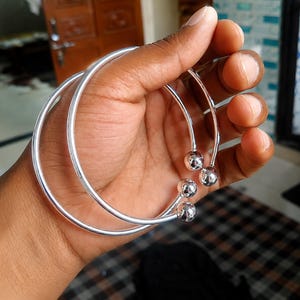 May include: Two polished silver-tone open cuff bracelets, each featuring a smooth, thin circular band that terminates at both ends with a small, spherical ball bead. The bracelets have a reflective, metallic finish and a simple, open-ended design.