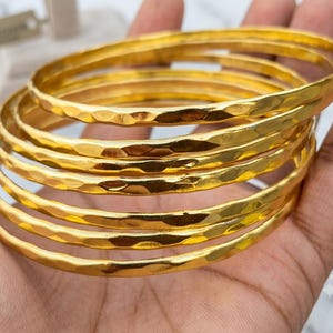 May include: A set of five gold-colored bangles featuring a hammered, textured surface. Each bangle is circular and reflects light across its uneven, metallic finish, creating a stacked appearance when held together in a group.