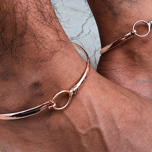 May include: A pair of polished copper-toned metal anklets, each featuring a smooth, rigid band design that connects to a circular ring clasp. The jewelry has a reflective, metallic finish and includes the text PURVIJEWELLER printed on the side.