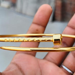 Könnte beinhalten: Ein goldfarbener Metallarmreif in Form eines gebogenen Nagels. Das Armband besteht aus einem glatten, geschwungenen Schaft, der an einem Ende spitz zuläuft und am anderen in einem flachen, zylindrischen Nagelkopf endet, mit strukturierten Kerben.