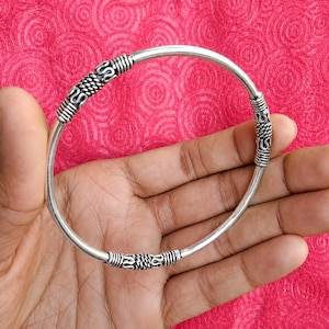 May include: A silver-toned metal bangle bracelet featuring a smooth, thin circular band accented with three distinct, textured cylindrical segments. Each segment displays a detailed, repeating pattern of horizontal ridges and twisted rope-like coils.