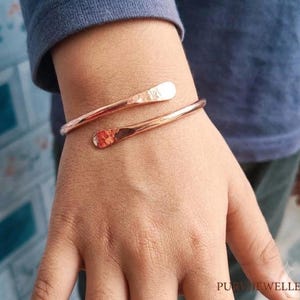 Könnte beinhalten: Ein kupferfarbenes Armband in Wickeloptik mit poliertem, gehämmertem Finish an den abgerundeten Enden. Das Metallband hat eine glatte, reflektierende Oberfläche, die sich um das Handgelenk legt und ein offenes Design mit zulaufenden Spitzen zeigt.