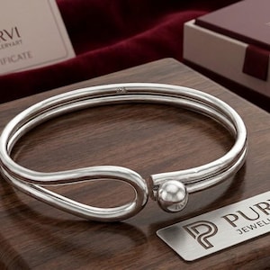 May include: A polished silver cuff bracelet featuring a double-band design that loops at one end and terminates with a smooth spherical bead at the other. The bracelet is displayed on a wooden surface with a Purvi Jewelleryart logo plate nearby.