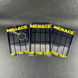 May include: Three black keychains with yellow text that reads "MENACE OF THE DAY". The keychains have a grid pattern and a silver keyring. The keychains are displayed on a gray surface.