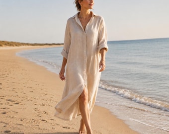 May include: A beige linen shirt dress with long sleeves and a button-down front. The dress falls to the ankles and is worn on a sandy beach with the ocean in the background. The dress is lightweight and flowing, suitable for warm weather.