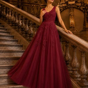 May include: A floor-length burgundy gown featuring a sleeveless bodice with a V-neckline, intricate lace floral appliques on the chest and waist, and a full, layered tulle skirt that flows downward from a fitted waistline.