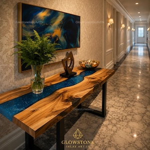 May include: A live edge wood console table with a central blue resin river inlay featuring embedded glitter. The table has black metal legs and holds a clear glass vase with green ferns, a dark abstract sculpture, and a gold bowl of spheres.