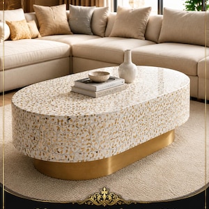 May include: An oval coffee table featuring a white surface with intricate gold-toned leaf-shaped inlay patterns. The table is supported by a solid, brushed gold-colored oval base. On top of the table sit two stacked books and a small ceramic vase.