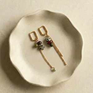 May include: Gold-toned earrings with rectangular hoops and dangling chains. Each earring features a cluster of beads in various colors, including black, white, green, and red. The earrings are displayed on a white, scalloped-edge plate.