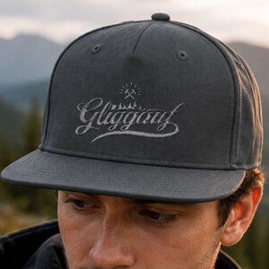 Könnte beinhalten: Eine anthrazitfarbene Five-Panel-Snapback-Cap mit dem in weiß gestickten Schriftzug Gliggauf in Schreibschrift, platziert über einer kleinen Grafik aus zwei gekreuzten Hämmern und einer Bergsilhouette auf der Vorderseite.