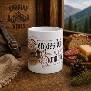 Könnte beinhalten: Ein weißer Keramikbecher mit einer dekorativen rot-schwarzen Schrift mit dem Text Vergass dei Samit nei, ergänzt durch eine kleine Illustration einer Berglandschaft mit Tannen, platziert auf einer Holzoberfläche neben Lebensmitteln.