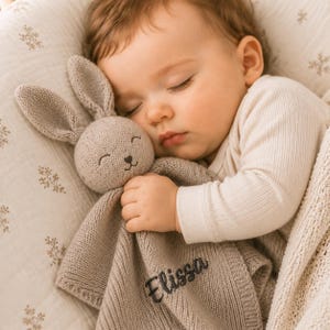 May include: A light beige knitted security blanket featuring a plush rabbit head with embroidered facial features and the name Elissa stitched in dark thread on the corner. The soft fabric has a textured knit pattern and a simple border edge.