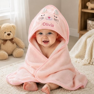 May include: A soft, light pink hooded baby towel featuring an embroidered cat face on the hood with closed eyes, whiskers, and a pink nose. The name Olivia is embroidered in pink script below the cat face. The towel has a plush, fuzzy texture.