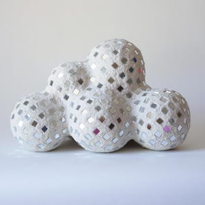 May include: A cloud-shaped decorative object covered in small, square mirrored tiles. The sculpture is white with silver and pink accents from the mirrored tiles. The cloud has a textured surface and is likely made of paper mache.