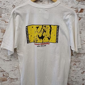 May include: A white short-sleeve t-shirt featuring a rectangular graphic on the back. The graphic depicts a black illustration on a yellow background framed by film strips, with the red text Princess MONONOKE and black text WILL TO LIVE.