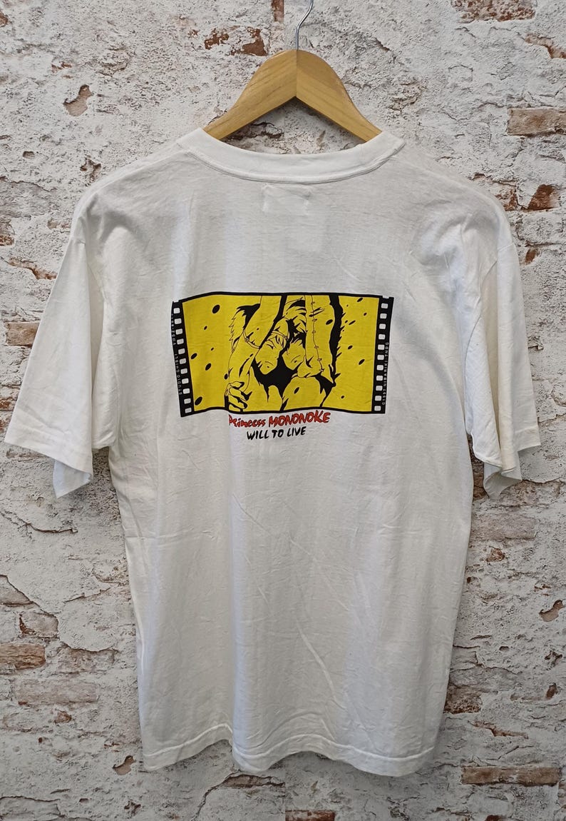 May include: A white short-sleeve t-shirt featuring a rectangular graphic on the back. The graphic depicts a black illustration on a yellow background framed by film strips, with the red text Princess MONONOKE and black text WILL TO LIVE.