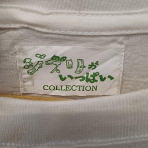 May include: A white t-shirt featuring a rectangular fabric label sewn into the inner neckline. The label displays green Japanese text and line art illustrations of small figures, with the word COLLECTION printed in green capital letters below.