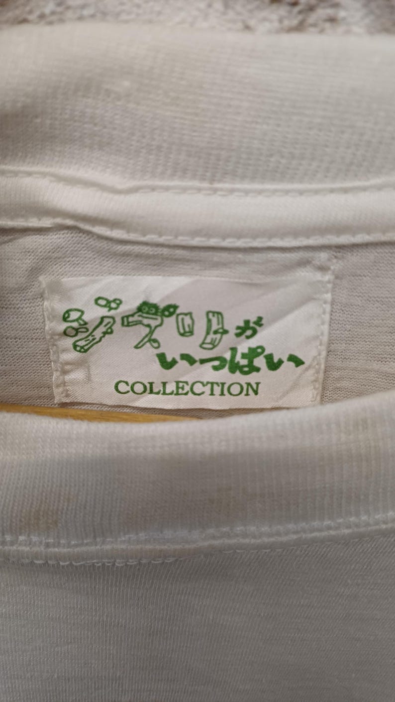 May include: A white t-shirt featuring a rectangular fabric label sewn into the inner neckline. The label displays green Japanese text and line art illustrations of small figures, with the word COLLECTION printed in green capital letters below.