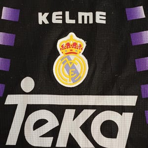 May include: A black sports jersey featuring purple horizontal stripes on the sides, a white collar, and the text Kelme and Teka. The chest displays a Real Madrid crest with a crown, and the inner neck label shows the text Ponedcloting:) and a paw print.
