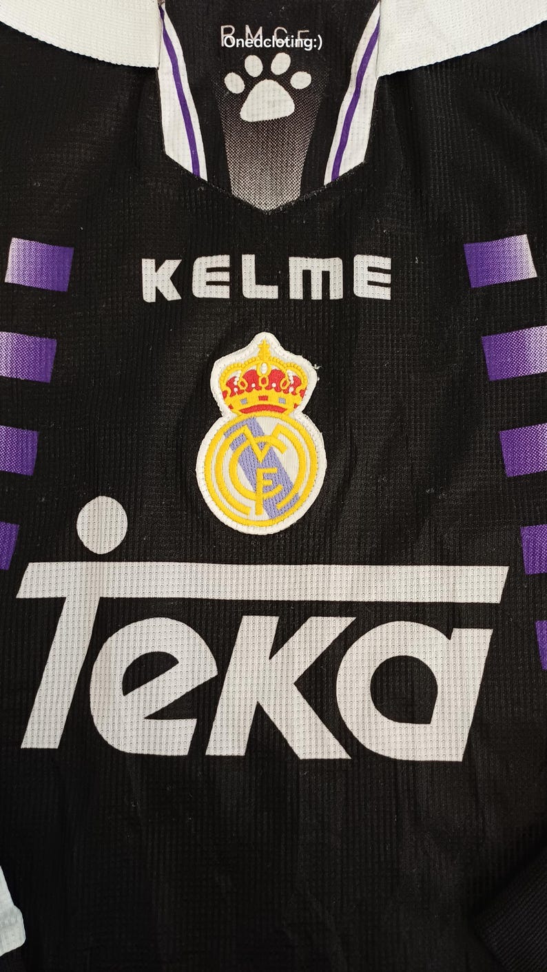 May include: A black sports jersey featuring purple horizontal stripes on the sides, a white collar, and the text Kelme and Teka. The chest displays a Real Madrid crest with a crown, and the inner neck label shows the text Ponedcloting:) and a paw print.