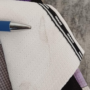 May include: A white fabric panel with a textured surface and a black striped border featuring the text Onedcloting:). A blue pen points to the fabric, which is layered over a black and purple patterned textile. The fabric has a visible weave.