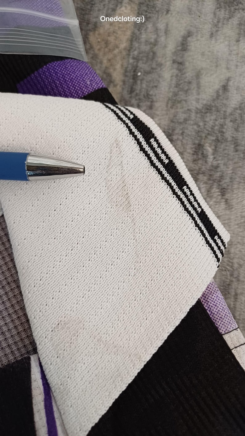 May include: A white fabric panel with a textured surface and a black striped border featuring the text Onedcloting:). A blue pen points to the fabric, which is layered over a black and purple patterned textile. The fabric has a visible weave.