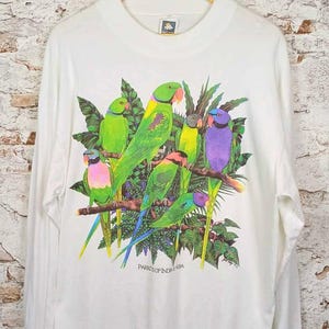 Puede incluir: Una camiseta blanca de manga larga con un estampado gráfico colorido de varios loros posados en ramas entre follaje verde. El texto "PARROTS OF INDONESIA" está impreso en negro debajo de las aves. La camiseta tiene cuello redondo.